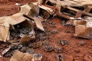 Damaged cardboard boxes and rusted ball bearings scattered on red soil after pallet collapse